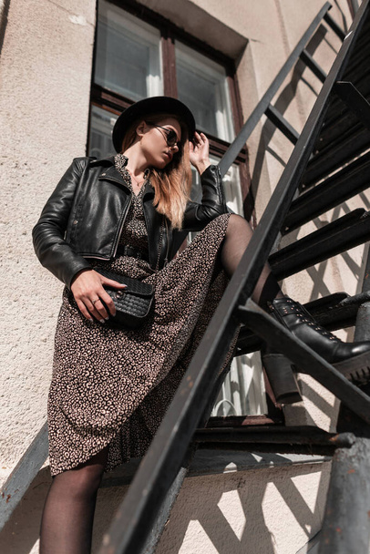 Elegáns gyönyörű modell lány kalapban, napszemüvegben, divatos ruhában, bőrdzsekivel és fekete kézitáskával egy fém lépcsőn áll a városban. - Fotó, kép