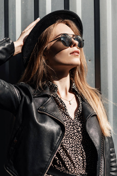 Fashion portrait of beautiful young woman with black glasses and fashionable hat with vintage dress and casual leather jacket outdoors on a sunny day - Fotoğraf, Görsel