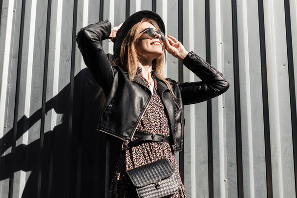 Menina elegante bonita feliz com óculos de sol estilo em vestido vintage com jaqueta de couro preto e bolsa desfrutar perto de uma parede de metal - Foto, Imagem