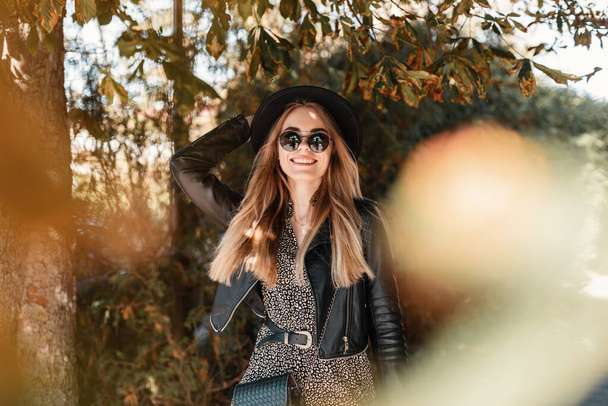 Happy portrait of a beautiful hipster girl with sunglasses in fashion clothes with a leather jacket and a hat are walking near the autumn foliage on the street - Photo, Image