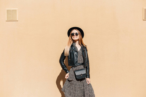 Happy beautiful young woman model in trendy casual look clothes with a hat and small black handbag stands near a beige wall on a sunny day. Feminine style, fashion and beauty - Photo, Image