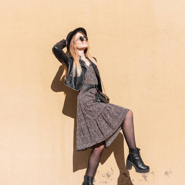 Забавный портрет красивой счастливой женщины в солнечных очках в модном наряде с платьем, кожаной курткой и туфлями, позирующими возле бежевой стены. Стиль и красота моды - Фото, изображение