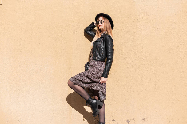 Hübsche junge sinnliche Frau im Hipster-Outfit mit Jacke, Kleid, Sonnenbrille, Hut und Schuhen posiert in der Nähe einer Wand. - Foto, Bild