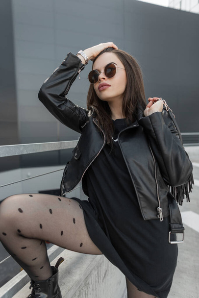 Beautiful sexy model woman with stylish round sunglasses in a fashionable leather jacket and black sweatshirt with sexy tights holds a bag and posing on the street - Photo, image