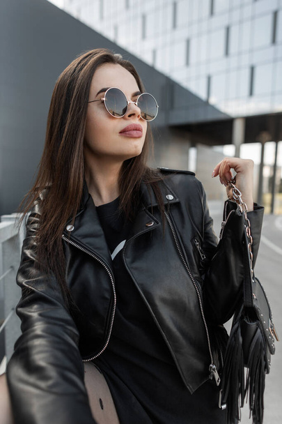 Fashion stylish beautiful model woman in vintage round sunglasses with fashionable leather jacket and black dress with stylish handbag walks in the city - Foto, Imagem