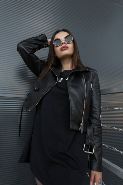 Mooie stijlvolle vrouw draagt zwarte leren jas loopt in de buurt van een metalen muur in de stad. Kleding, stijl en mode - Foto, afbeelding