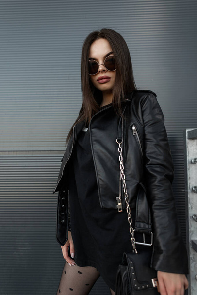 Havalı güneş gözlüklü, siyah elbiseli, siyah ceketli ve şehirde metal bir duvarın yanında şık bir çantası olan güzel bir genç kadın. Kopyala, metin için boşluk - Fotoğraf, Görsel