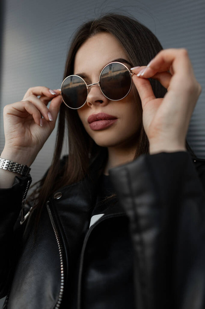 Fashion cool portrait of a beautiful hipster woman with big lips in stylish black leather jacket puts on fashionable round sunglasses in the city - Foto, Bild