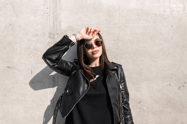 Klasik güneş gözlüklü ve deri ceketli, modaya uygun siyah elbiseli doğal güzelliğe sahip bir kadın model şehirde güneş ışığında beton bir duvarın yanında duruyor. - Fotoğraf, Görsel