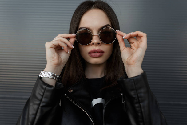 Fashion portrait of a beautiful hipster woman model with big natural lips in a stylish black leather jacket puts up cool round sunglasses near the metal wall on the street - Foto, immagini