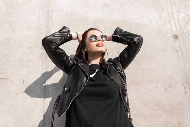 Retrato de moda de una hermosa mujer modelo en ropa de cuero negro elegante con gafas de sol vintage posando cerca de una pared de hormigón gris en un clima soleado - Foto, imagen