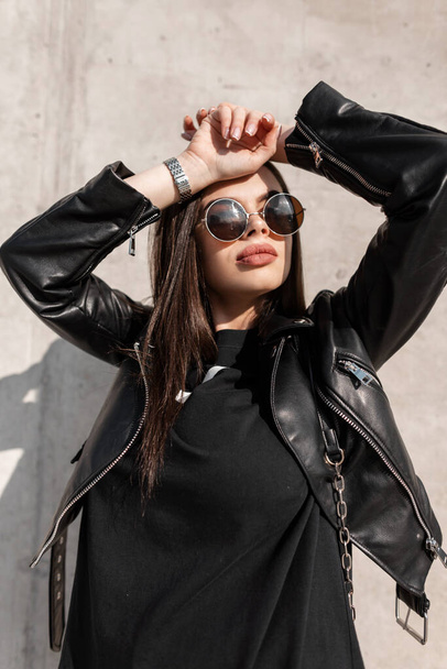 Stijlvolle mooie jonge vrouw met vintage ronde zonnebril in de mode zwart lederen jas en zwart staat in de buurt van een grijze betonnen muur op een zonnige dag - Foto, afbeelding