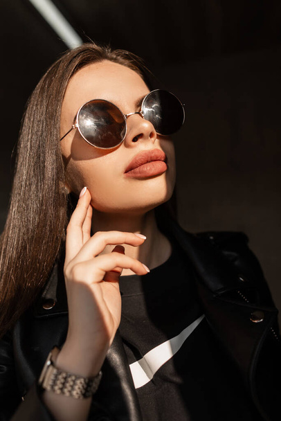 Stilvolles, kreatives Porträt einer jungen Frau mit natürlichem Gesicht und großen Lippen in schwarzer modischer Kleidung und Vintage-Sonnenbrille steht im Sonnenlicht und Schatten auf dunklem Hintergrund - Foto, Bild