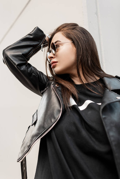 Hipster beautiful cool woman with trendy sunglasses in fashion rock leather jacket and black sweatshirt on gray background walks in the city. Urban portrait of a female model - Photo, Image
