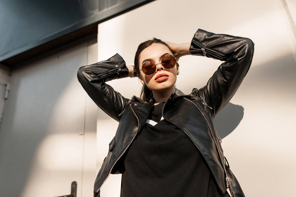 Trendy young beautiful woman model in fashion black clothes with leather stylish jacket with vintage round sunglasses walks in the city at sunset - Fotó, kép