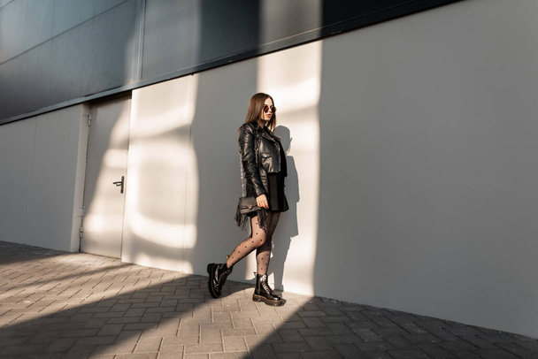Κομψό όμορφο δροσερό κορίτσι μοντέλο με vintage γυαλιά ηλίου σε μαύρα μοντέρνα ρούχα με δερμάτινο μπουφάν, φόρεμα, σέξι καλσόν και μπότες βόλτες στην πόλη στο φως του ήλιου - Φωτογραφία, εικόνα