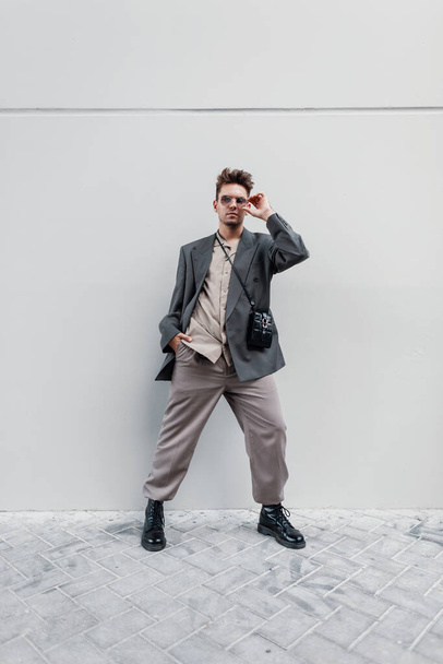 Κομψό όμορφο ανδρικό μοντέλο με γυαλιά ηλίου σε μοντέρνο σακάκι με πουκάμισο, τσάντα, παντελόνι και sneakers στέκεται σε γκρι φόντο - Φωτογραφία, εικόνα