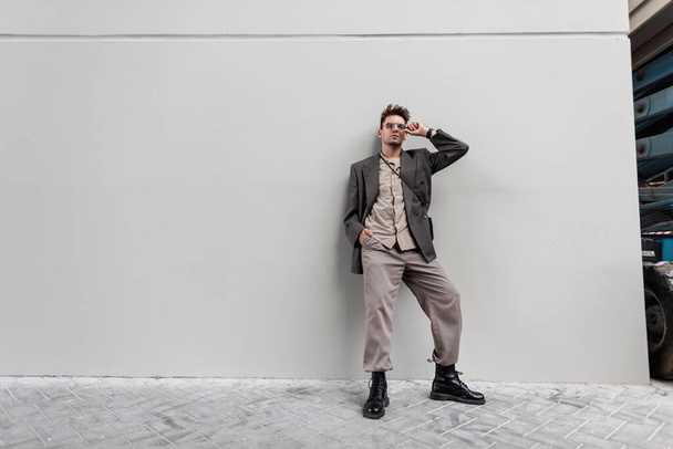 Modny młody człowiek w stylowym stroju casual wygląd z ponadgabarytowej marynarki, koszuli i czarnych butów stoi w pobliżu szarej ściany w mieście - Zdjęcie, obraz