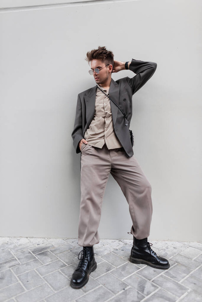 Όμορφος νεαρός άνδρας με γυαλιά ηλίου στη μόδα ντύσιμο φορούν σακάκι, πουκάμισο και δερμάτινα παπούτσια στέκεται κοντά γκρι τοίχο στο δρόμο. Αστική ανδρική μόδα - Φωτογραφία, εικόνα