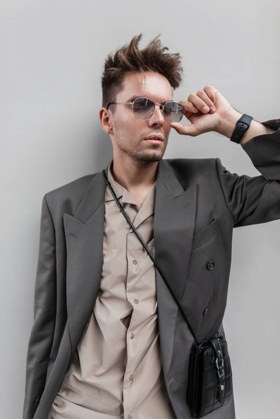 Stilvoller junger gutaussehender Mann mit Frisur und Bart in modischer Freizeitkleidung mit Blazer und Hemd mit Handtasche trägt Sonnenbrille in der Nähe der grauen Mauer in der Stadt - Foto, Bild