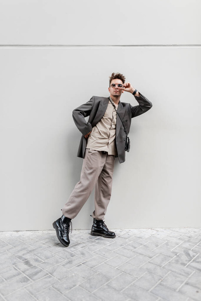 Trendy Geschäftsmann Kerl Modell in modischen Look Kleidung mit Sonnenbrille geht auf der Straße in der Nähe einer grauen Wand. Lässige Mode, Stil und Schönheit für Männer - Foto, Bild