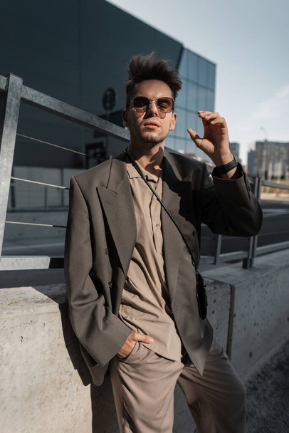 Fashion portrait of a beautiful young man model with stubble in a fashionable suit with a jacket and shirt puts on sunglasses in the city. Male urban business style - Photo, Image