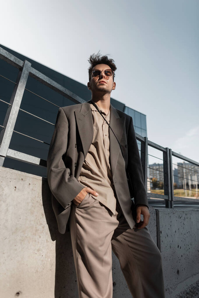 Stylový pohledný obchodník modelka v módním šedém obleku se slunečními brýlemi ve městě. Městský mužský styl - Fotografie, Obrázek