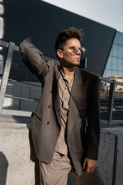 Stylish urban portrait of a handsome young guy with bristles in a fashionable suit with a shirt and a blazer in cool sunglasses stands on the city at sunlight - Фото, изображение
