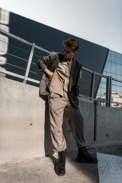 Cool modelo de hombre joven con gafas de sol vintage en blazer gris moda, camisa, pantalones y botas negras se encuentra en el fondo urbano a la luz del sol. Estilo casual de negocios - Foto, imagen