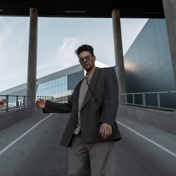Stijlvolle knappe jonge man met modieuze bril en kapsel in casual kleding met een blazer, shirt en broek wandelingen in de stad tegen de achtergrond van een modern gebouw - Foto, afbeelding
