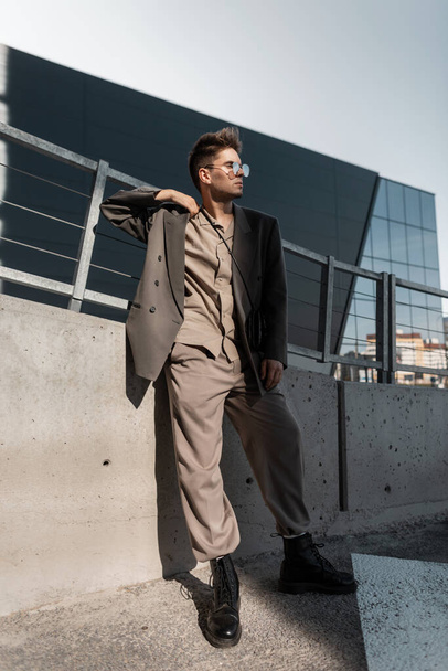 Handsome young business man model with stylish sunglasses in fashionable gray suit stands in the city at sunlight. Male urban elegant style, fashion and success - Фото, изображение