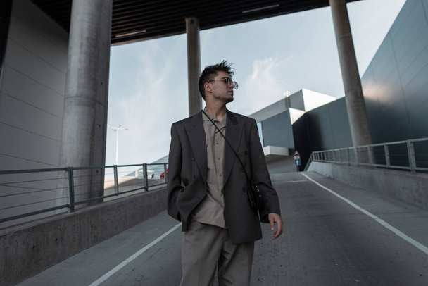 Bonito homem de negócios jovem com óculos de sol em um terno cinza na moda com uma bolsa de mão caminha na cidade perto de um edifício moderno - Foto, Imagem