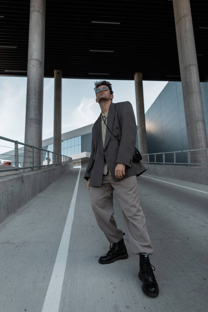 Elegante joven guapo en traje gris de moda con chaqueta, camisa, botas y gafas de sol en la ciudad. Estilo urbano masculino y belleza - Foto, Imagen