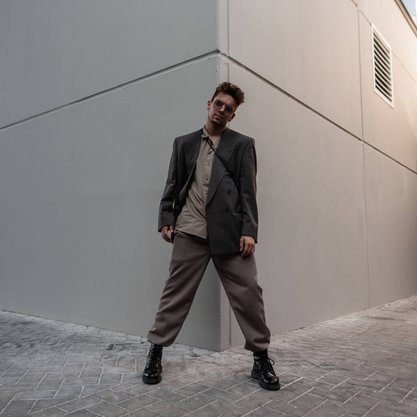 Красивый модный мужчина модель в стильном сером костюме с модными солнцезащитными очками стоит рядом с современным зданием в городе - Фото, изображение