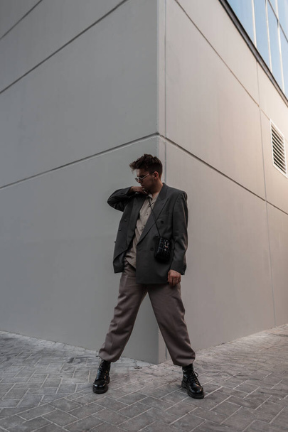 hombre hipster guapo de moda en ropa de aspecto elegante con zapatos y gafas de sol con estilo se encuentra cerca de la pared gris en la ciudad. Estilo urbano masculino y moda - Foto, imagen