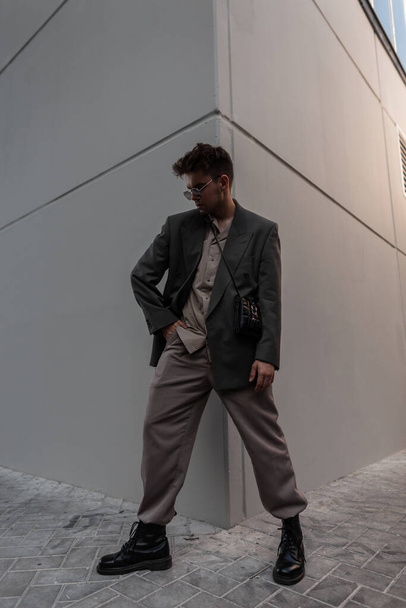 Cool handsome hipster man with glasses in fashionable clothes with jacket, shirt, trousers and shoes near gray background in city - Foto, afbeelding