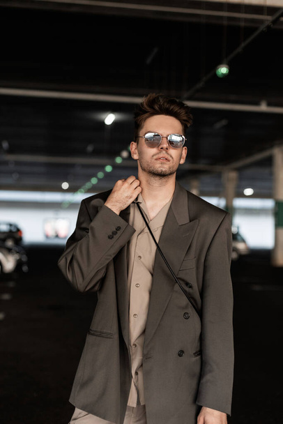 Stylowy mężczyzna biznesmen model z okularami przeciwsłonecznymi w marynarce mody i koszula spacer na parkingu - Zdjęcie, obraz