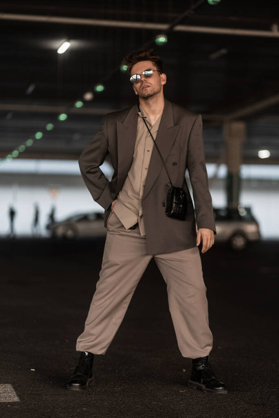 módní hezký mladý cool muž v módním šedém obleku se slunečními brýlemi a kabelkou stojí na parkovišti - Fotografie, Obrázek