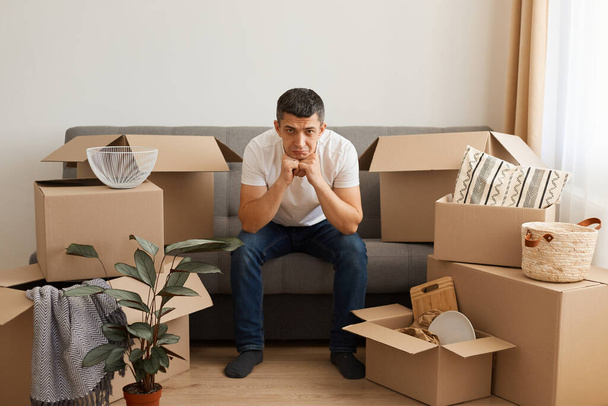 Indoor shot of sad upset man wearing white T-shirt and jeans sitting on sofa surrounded with cardboard boxes, looking at camera, expressing sorrow, doesn't want to move another apartment. - Foto, Imagem