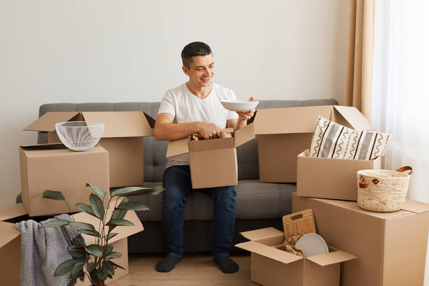 Indoor shot of positive optimistic man wearing white T-shirt and jeans sitting on sofa surrounded with cardboard boxes, unpacking packages, moving in a new house. - Valokuva, kuva