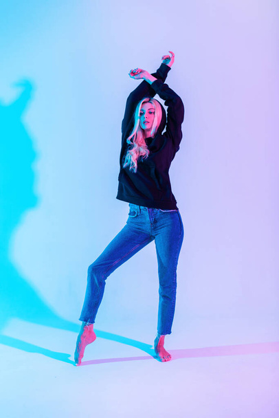 Elegante bella giovane donna in una felpa con cappuccio nera di moda con blu classici jeans danza su uno sfondo luminoso al neon rosa chiaro - Foto, immagini