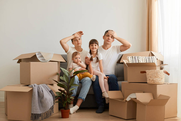 Portrait of tired couple sitting on sofa with children after moving, keeping hands on heads, exhausted of unpacking packages, expressing sorrow and fatigue. - Φωτογραφία, εικόνα