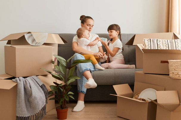 Pretty young mother and her cute little daughters sitting on cough of new apartment and unpacking moving boxes, expressing positive emotions, surrounded with packages with belongings. - Φωτογραφία, εικόνα