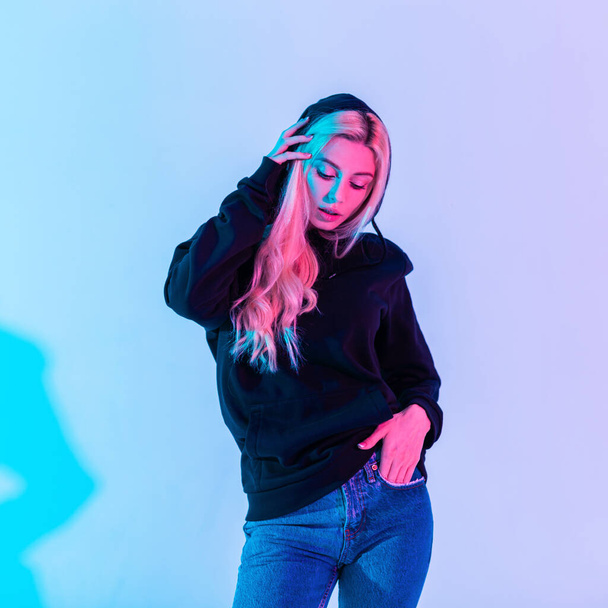 Colorful fashion portrait of beautiful stylish blonde woman in black sweatshirt wear a hood with blue jeans on pink and neon light - Photo, Image