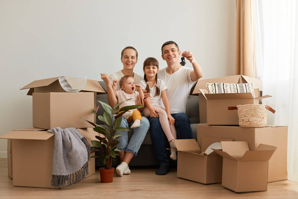 Portrait of extremely happy couple sitting on sofa with children after moving, buying new apartment, showing keys of house they are relocating, surrounded with cardboard boxes. - Foto, Bild