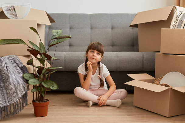 Portrait of pensive little girl wearing casual style attire sitting on a floor near sofa, holding chin, thinking about new room decor, having thoughtful facial expression, relocating. - Foto, afbeelding