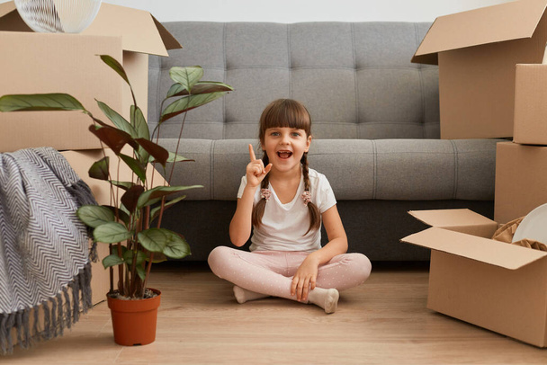 Portrait of positive excited little girl wearing casual style attire sitting on a floor with raised finger, pointing up, having idea of new room decor, expressing positive emotions. - Foto, imagen