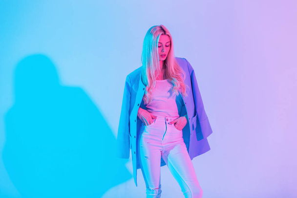 Kreatives, farbenfrohes Porträt eines jungen, schönen Mädchens in modisch eleganter Kleidung mit blauem Mantel, weißer Bluse und Jeans im Studio vor neonrosa Licht - Foto, Bild