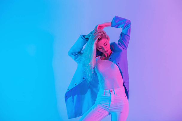 Glamorous stylish young blonde woman model in fashionable suit with blue blazer with white jeans poses on bright pastel colored neon pink lights in studio - Photo, Image