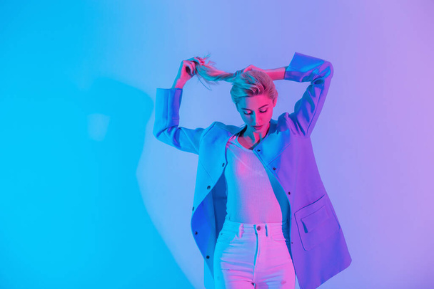 Retrato de moda colorido de bela jovem menina loira elegante na moda roupas olhar elegante com blazer azul, blusa branca e jeans em neon e rosa luz de fundo - Foto, Imagem